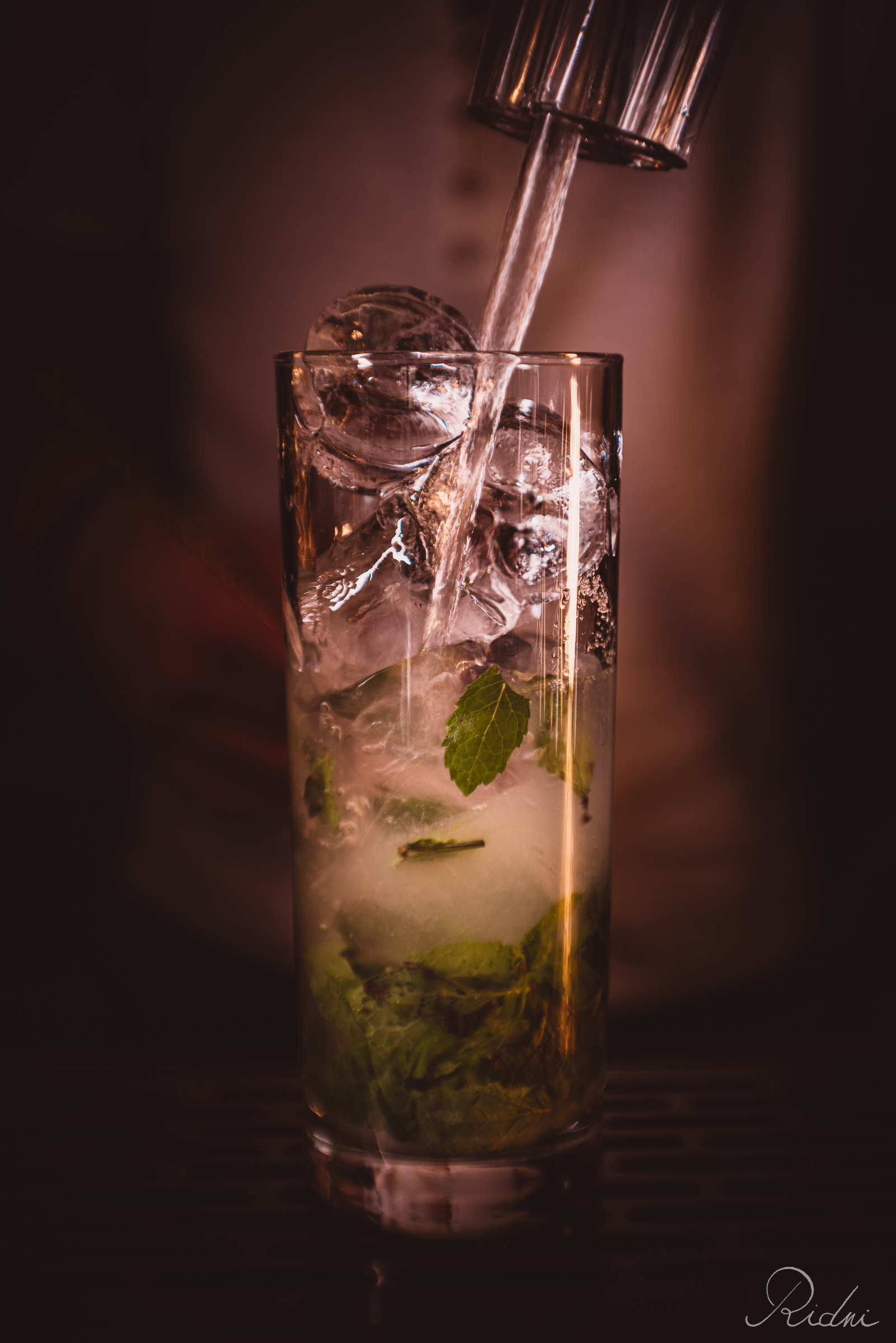 Liquid pours into a tall glass with mint and ice at bar Zona in lignano, friuli, highlighting expert hospitality in a moody setting.