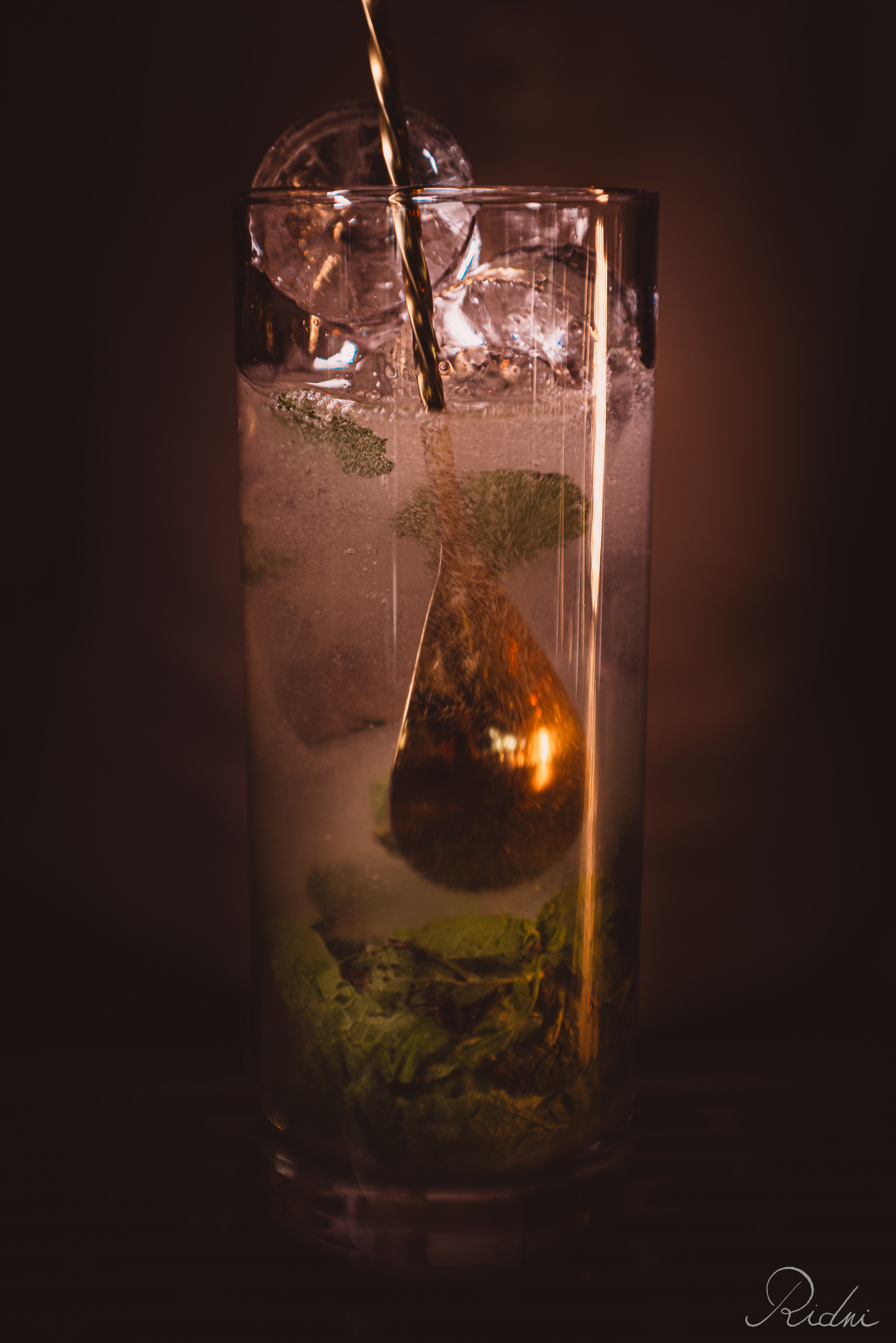 A golden bar spoon stirs a refreshing cocktail with mint and ice at bar Zona in lignano, friuli, captured in warm, moody lighting.