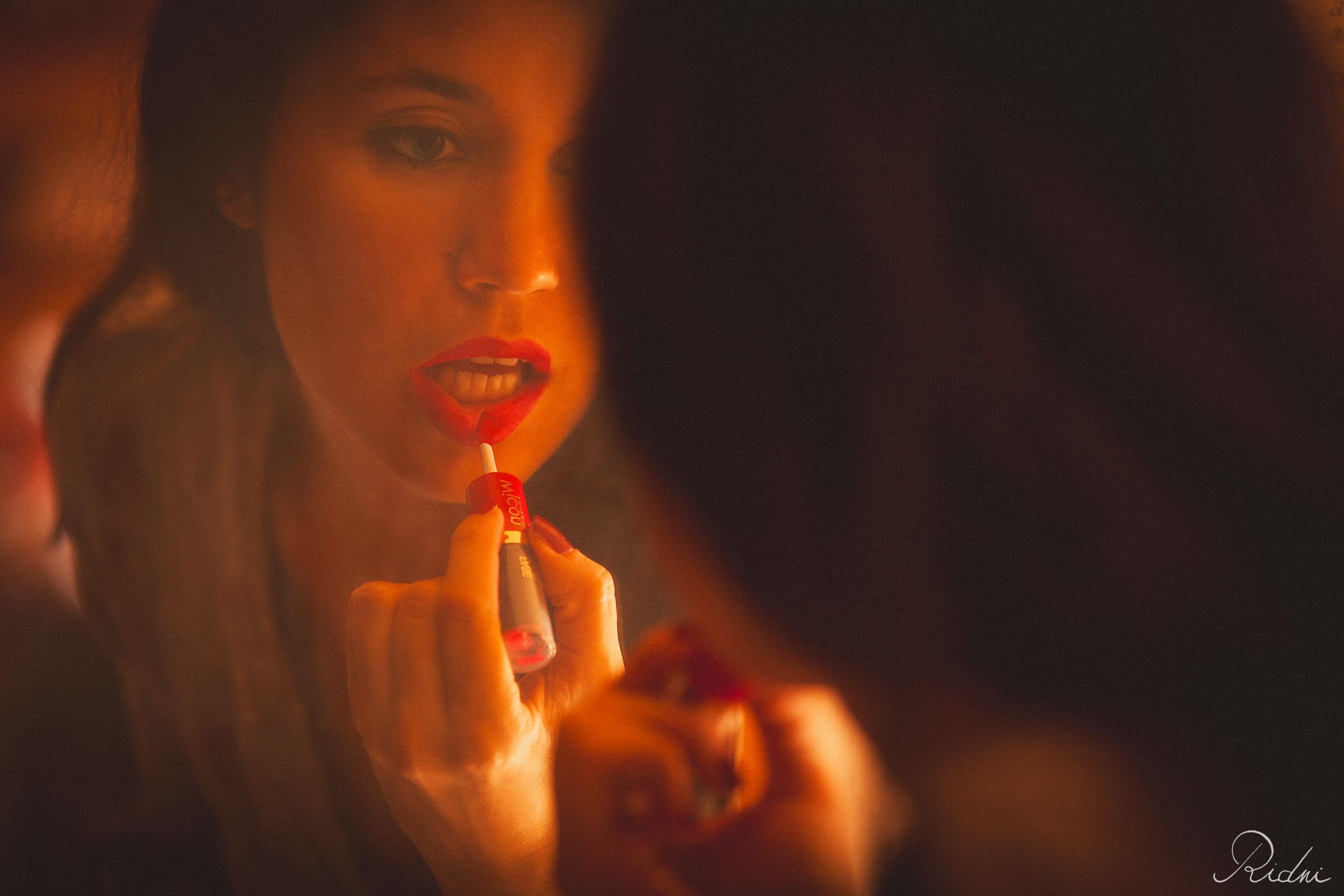 A woman applies vibrant red lipstick while looking into a mirror, captured in warm, cinematic lighting with a soft focus.