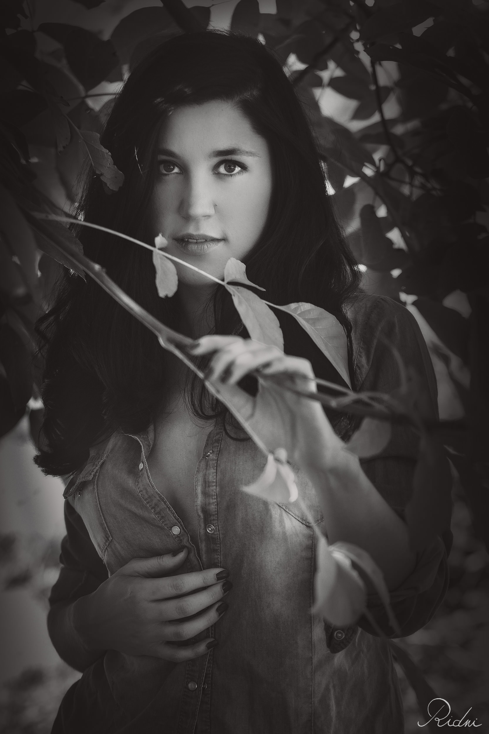 Black and white portrait of a woman with long dark hair, framed by leaves, wearing a denim shirt in a natural setting.