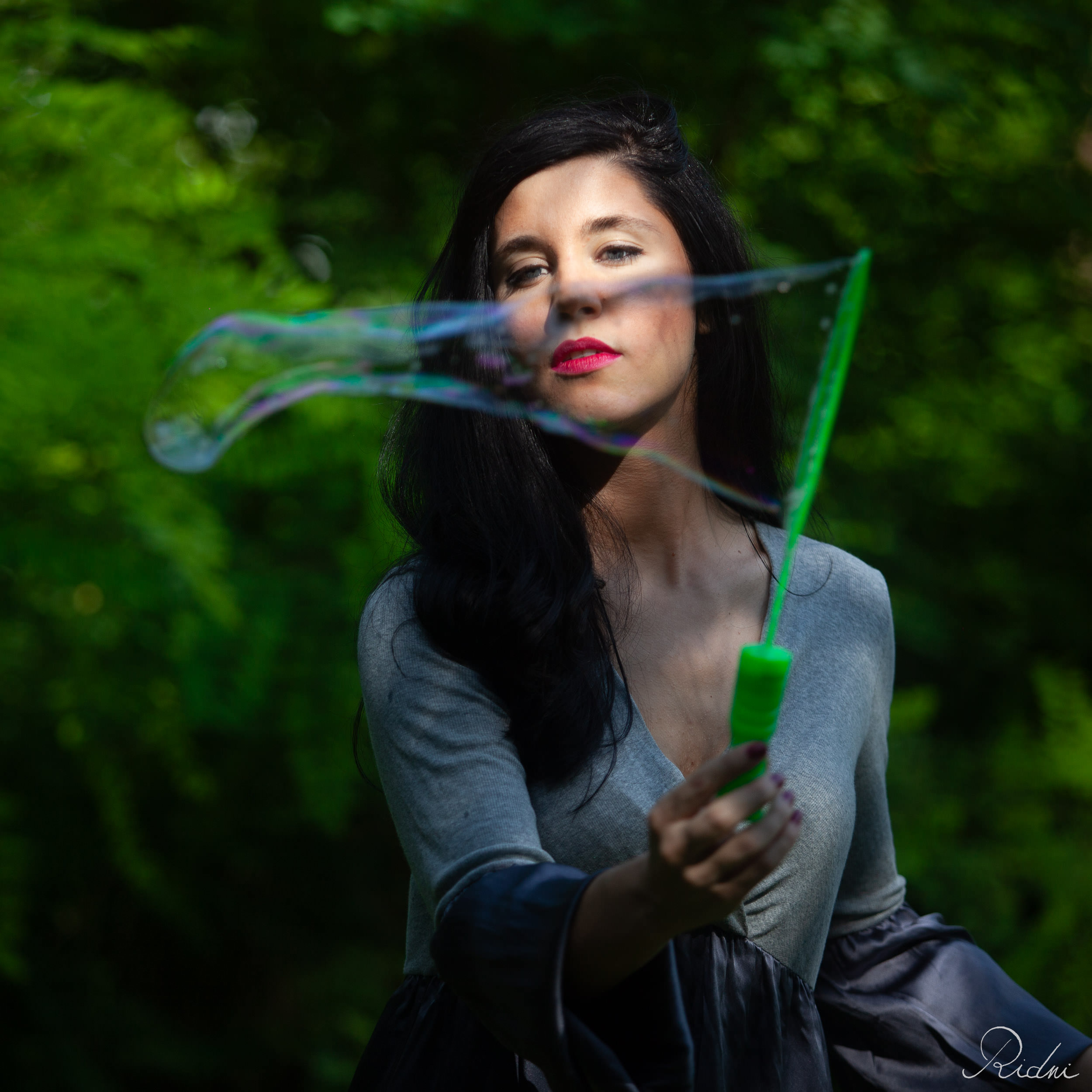 Dark-haired woman with red lipstick creates a large iridescent bubble with a green wand, set against a lush, sun-dappled forest background.