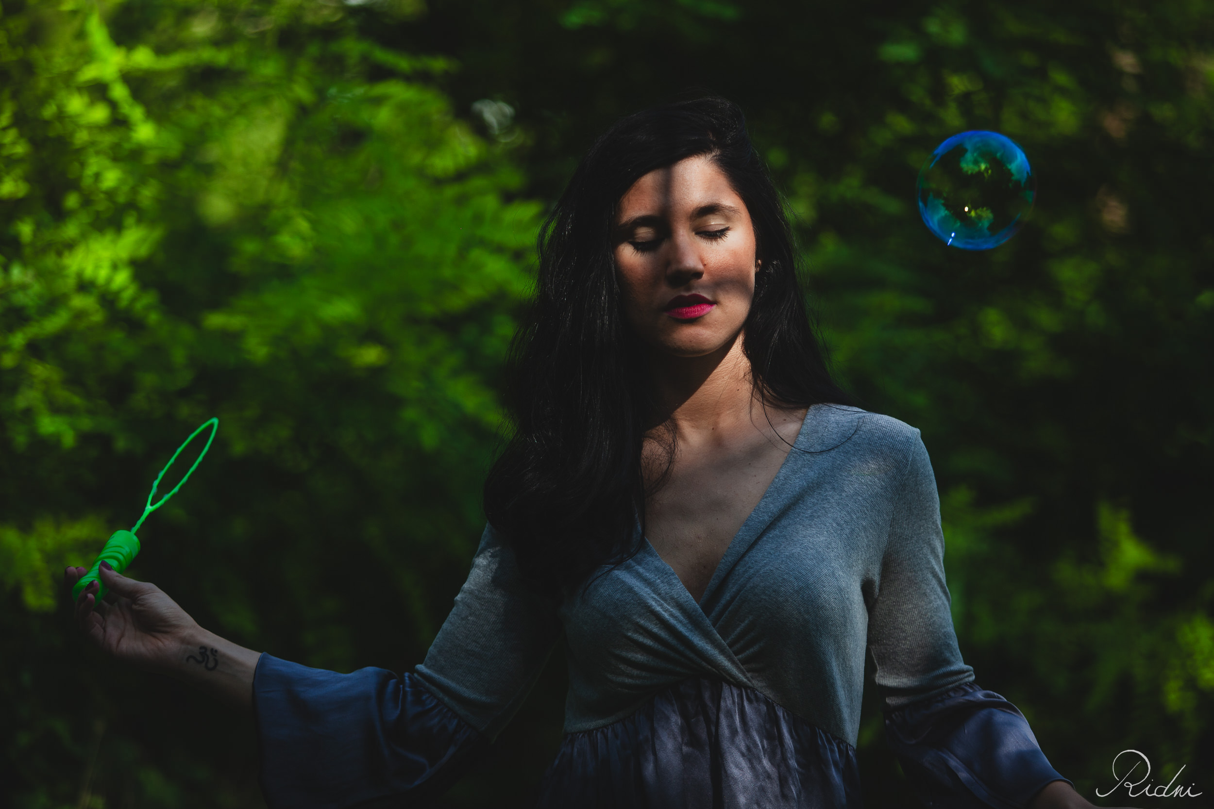 Serene woman with eyes closed stands amidst green foliage as a soap bubble floats by, captured in dramatic sunlight and shadow.