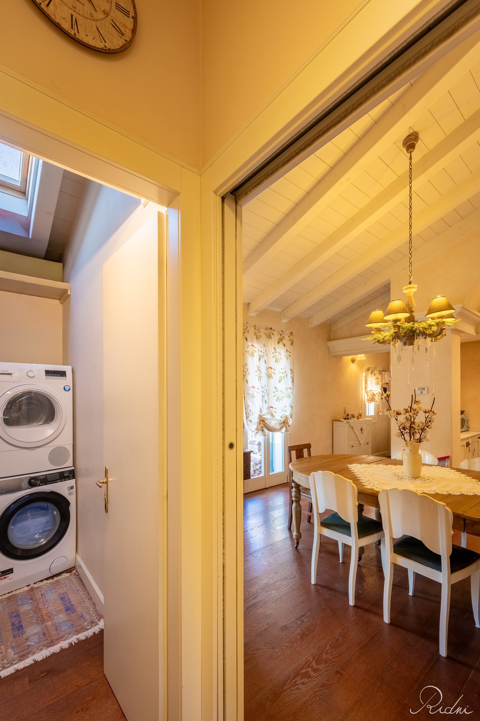 Stacked washer and dryer in a laundry nook beside a warm dining room with a chandelier, captured via real estate photography.