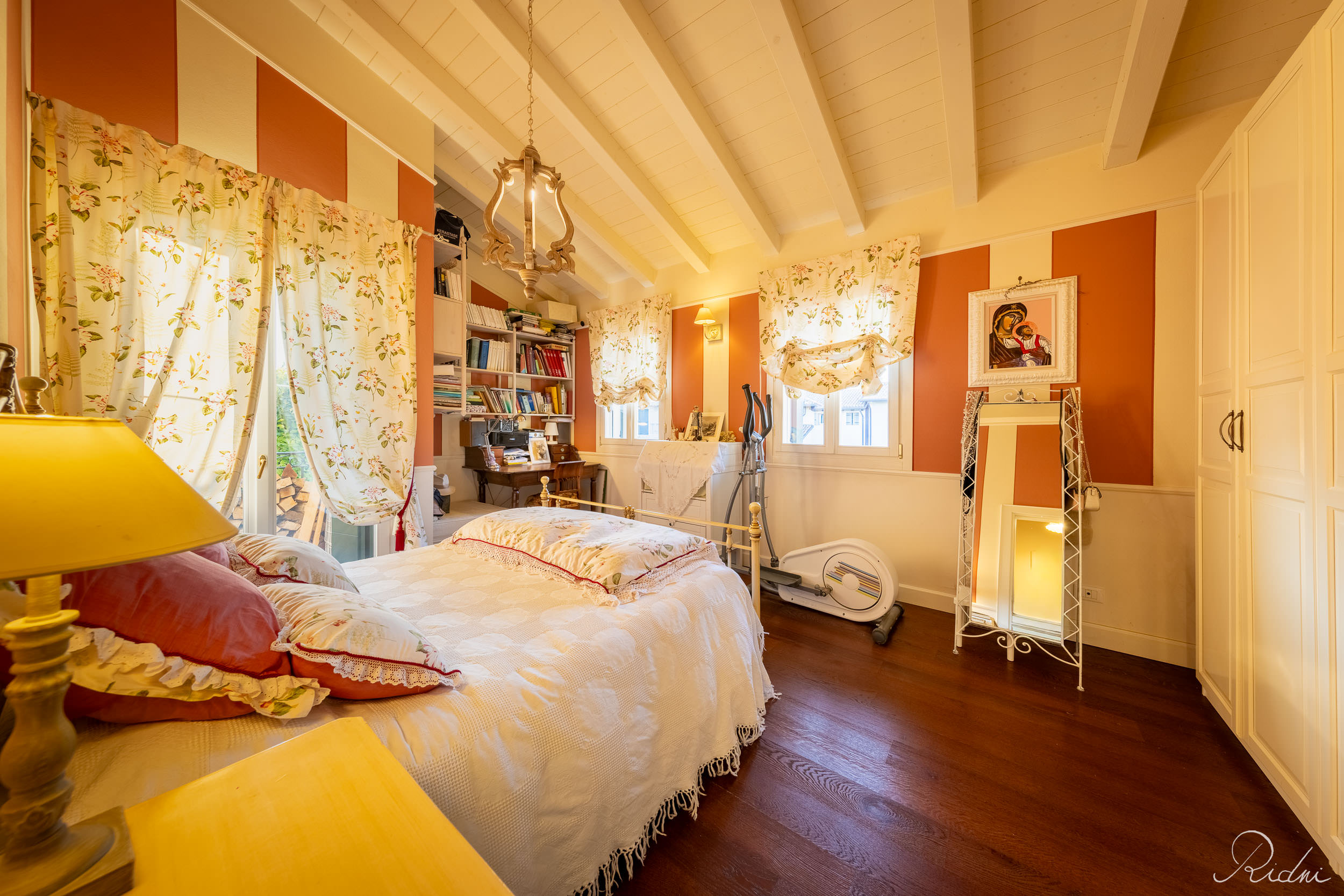 A cozy bedroom featuring sloped wooden ceilings, floral curtains, and orange striped walls, showcased through professional real estate photography.