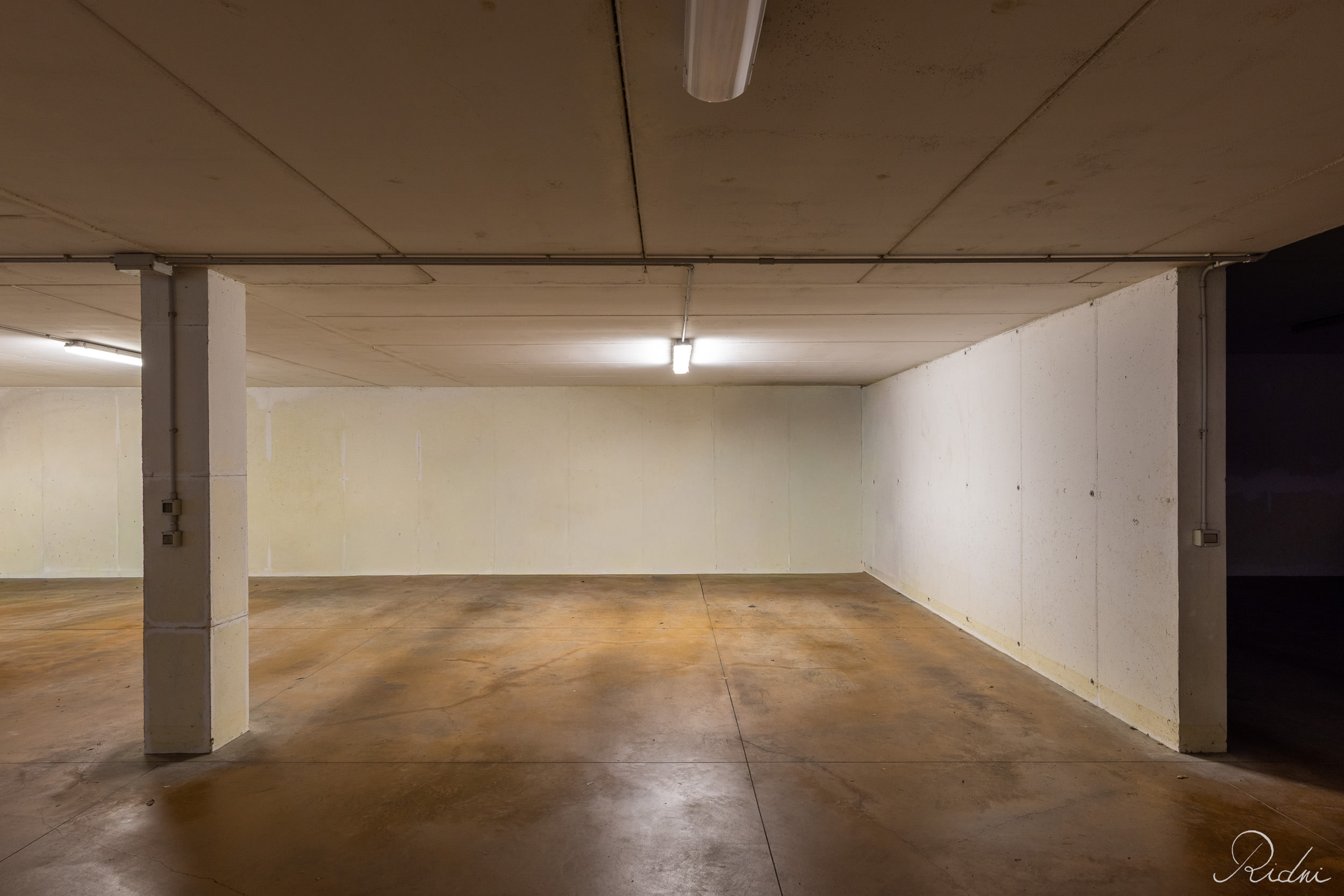 A spacious empty underground garage with concrete floors and fluorescent lighting, captured through professional real estate photography.